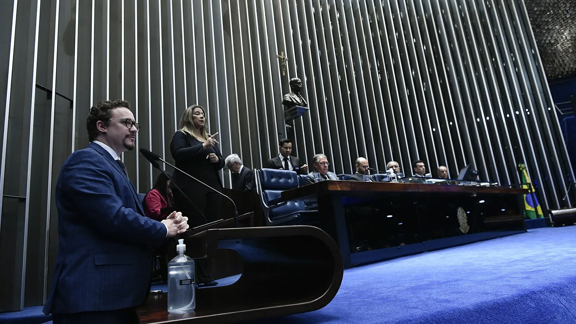 Plenário do Senado Federal, em Brasília, com orador à tribuna, intérprete de Libras e a Mesa diretora ao fundo, diante do painel de lâminas metálicas verticais.