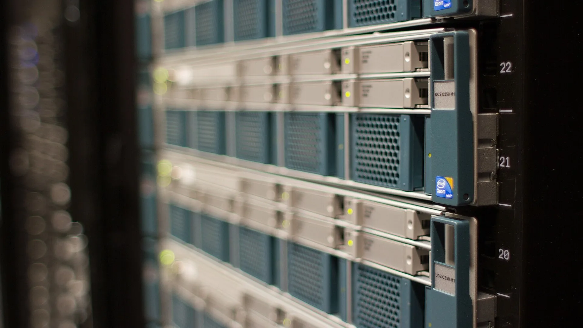 Fileiras de servidores montados em rack, com gavetas azul-petróleo e cinza e luzes de atividade desfocadas ao fundo.